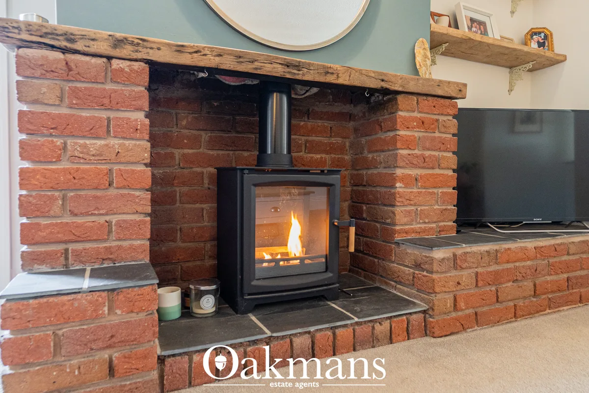 Contemporary wood-burning stove with active flame set within red brick fireplace alcove, wooden mantelpiece above, pale 