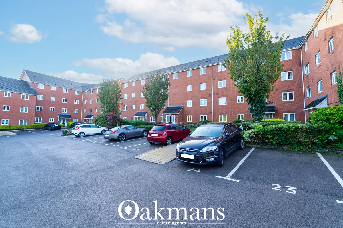 Red brick apartment building with parked cars in numbered spaces, landscaped grounds with shrubs and trees, clear blue s