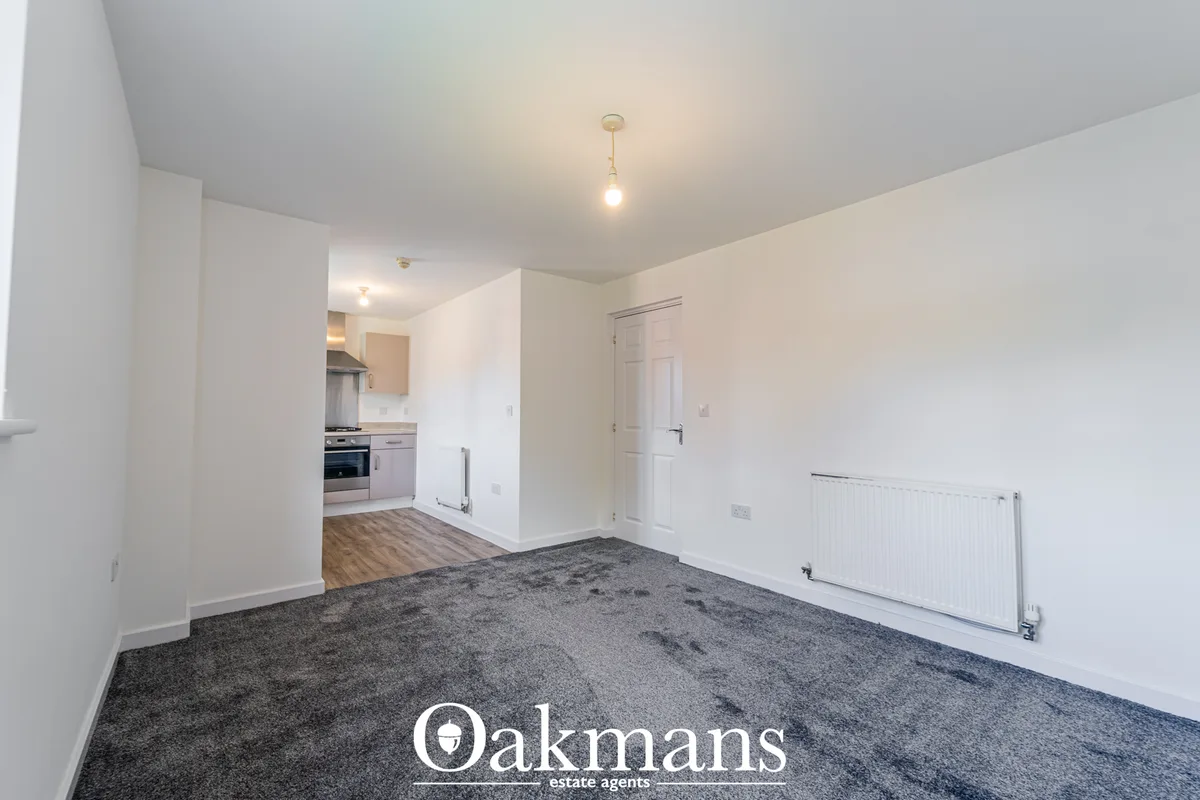 Open-plan living area with grey carpet, white walls, radiator, ceiling light, door to hallway, and kitchen visible beyon