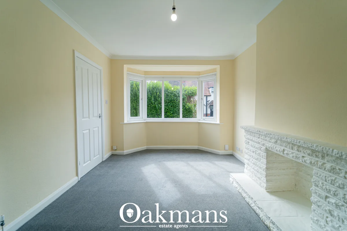 Bright hallway with cream walls, bay window overlooking green hedges, white panelled door, grey carpet and decorative br