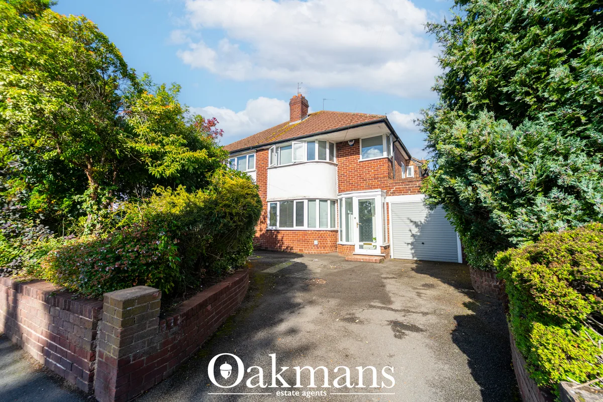 Red brick semi-detached house with white rendered upper section, bay windows, garage, and mature hedging either side of 