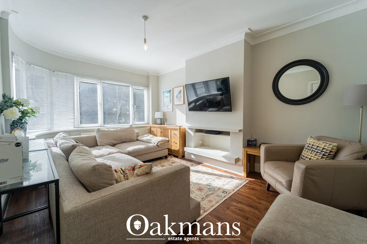 Spacious living room with beige L-shaped sofa, wall-mounted TV, wooden furniture, large windows with blinds, wooden floo