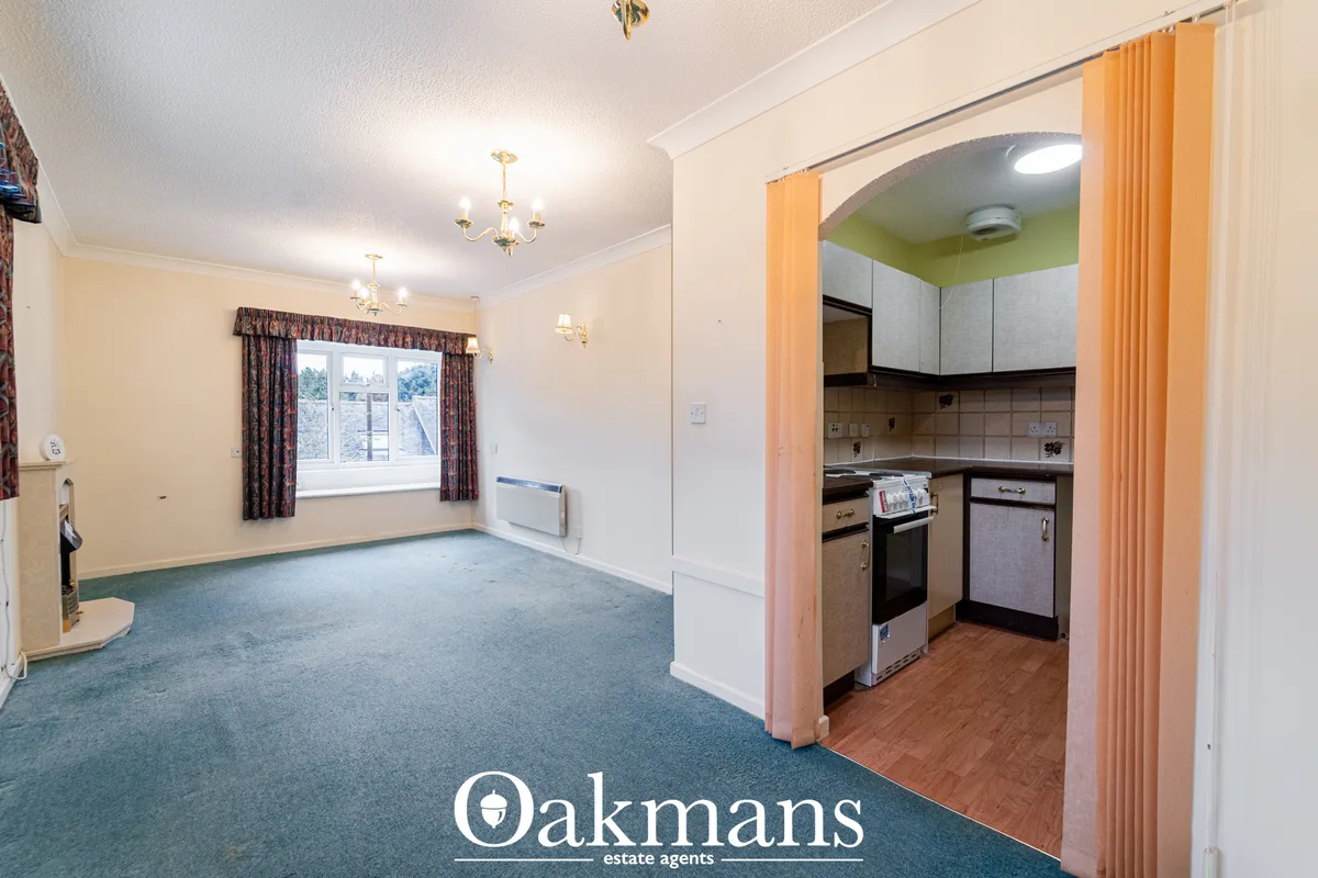 Spacious living room with cream walls, blue carpet, window with patterned curtains, wall lights, and open doorway to kit
