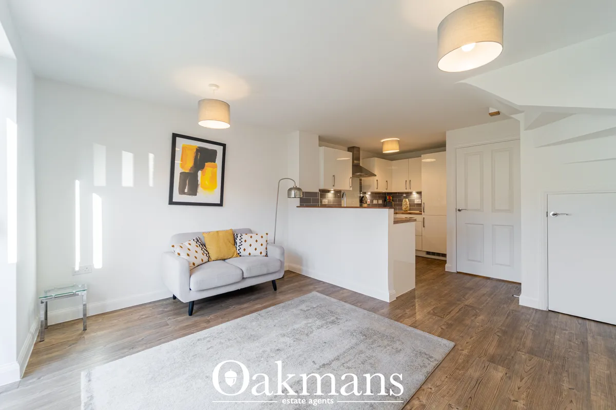 Open-plan living area with grey sofa, yellow accents, wooden flooring, white ceiling lights and open kitchen visible bey