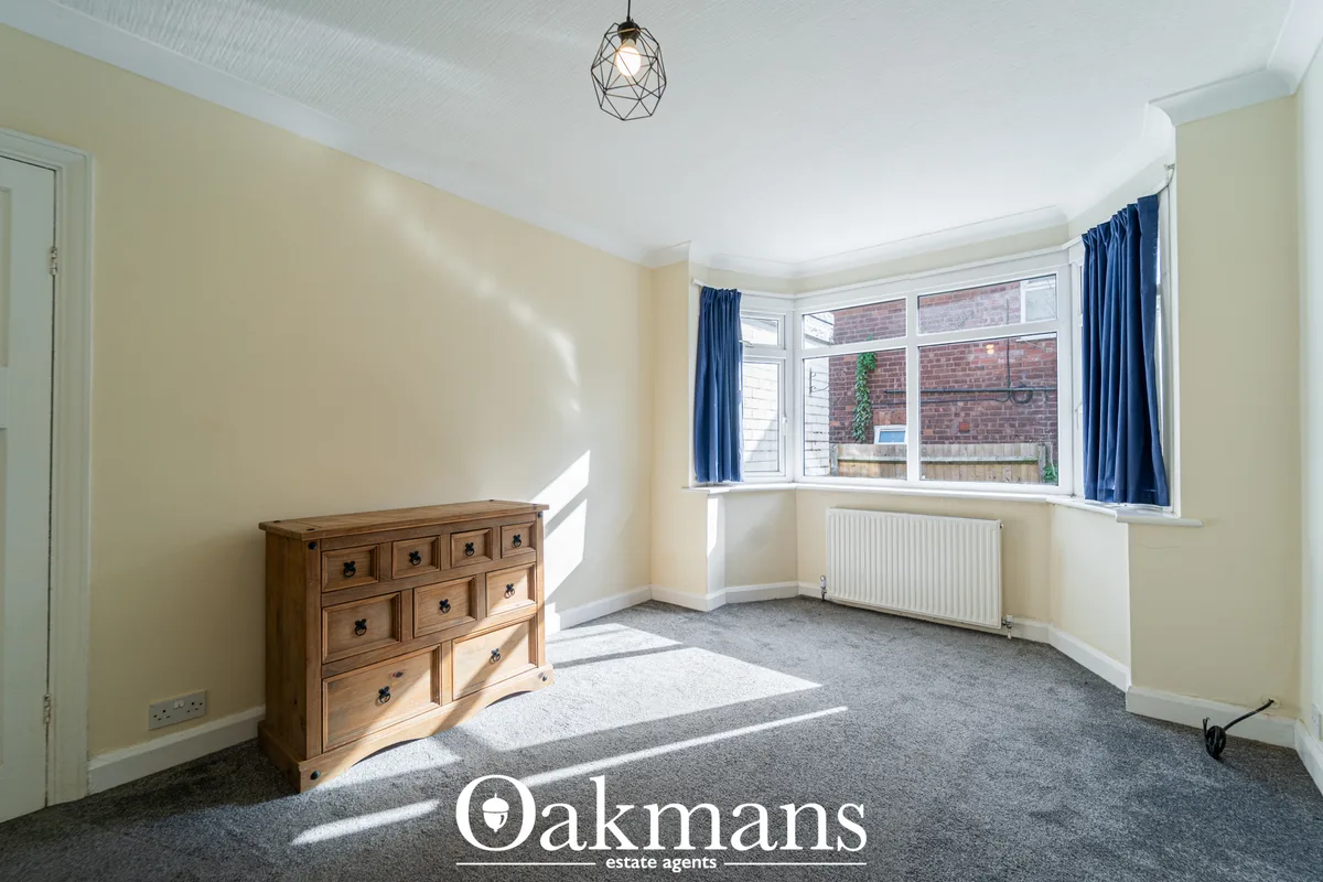 Bright living room with bay window, cream walls, grey carpet, wooden chest of drawers, blue curtains and geometric penda