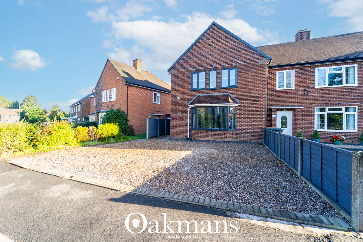 Red brick semi-detached house with gravel driveway, blue fence, multiple windows and pitched roof on sunny residential s