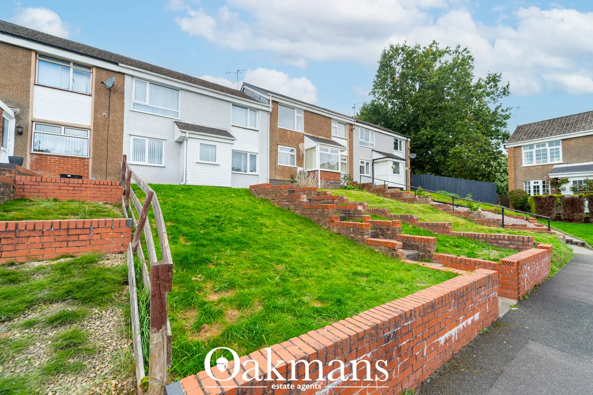 Terraced houses on sloped residential street with brick retaining walls, green lawns and mature trees under blue sky