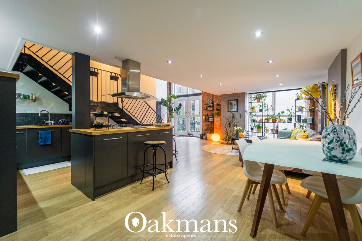 Open-plan living space with kitchen, dining area, staircase, wooden flooring, abundant plants, and floor-to-ceiling wind