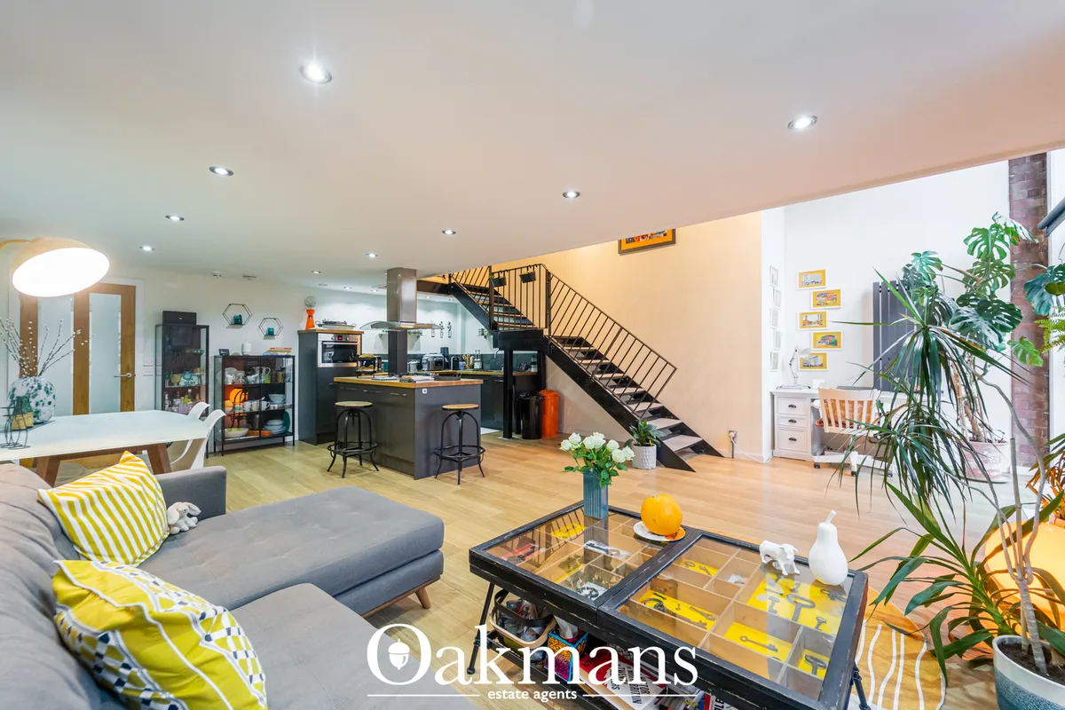 Open-plan living space with grey sofa, yellow striped cushions, wooden flooring, staircase, kitchen area and potted plan