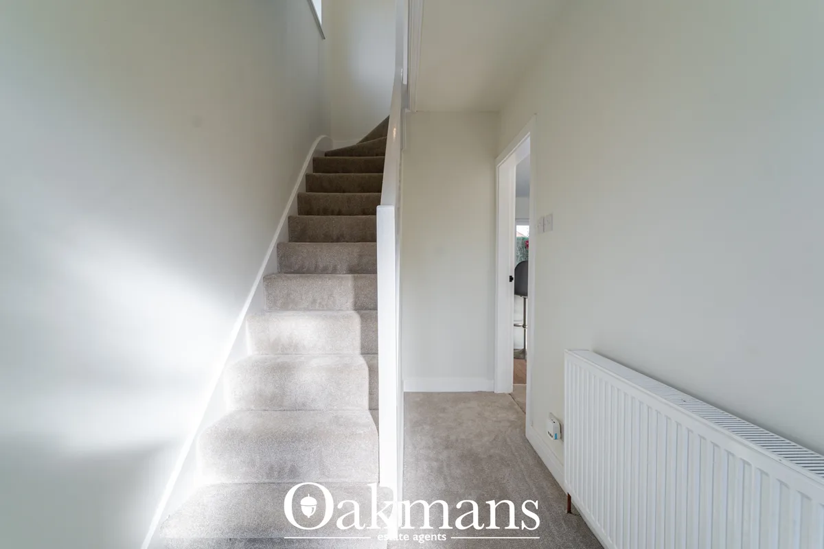 Bright hallway with staircase, white walls, grey carpet, radiator, and doorway to adjacent room