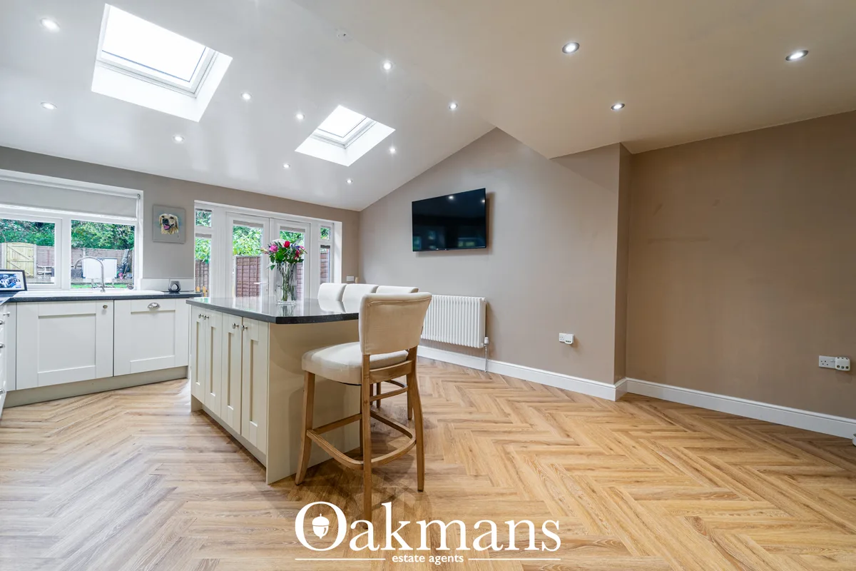 Open-plan living space with cream walls, skylight ceiling, herringbone wood flooring, wall-mounted TV, and breakfast bar