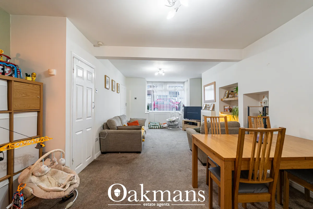 Open-plan living and dining area with grey sofa, wooden dining table, shelving unit, TV, large window with blinds and wo