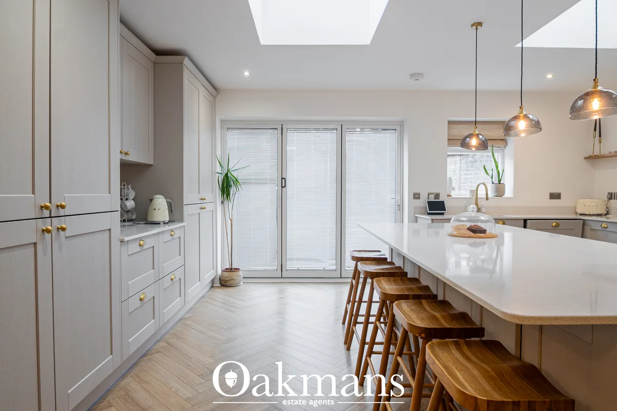 Modern fitted kitchen with white cabinetry, white worktops, bronze pendant lights, wooden bar stools, and bi-fold doors 