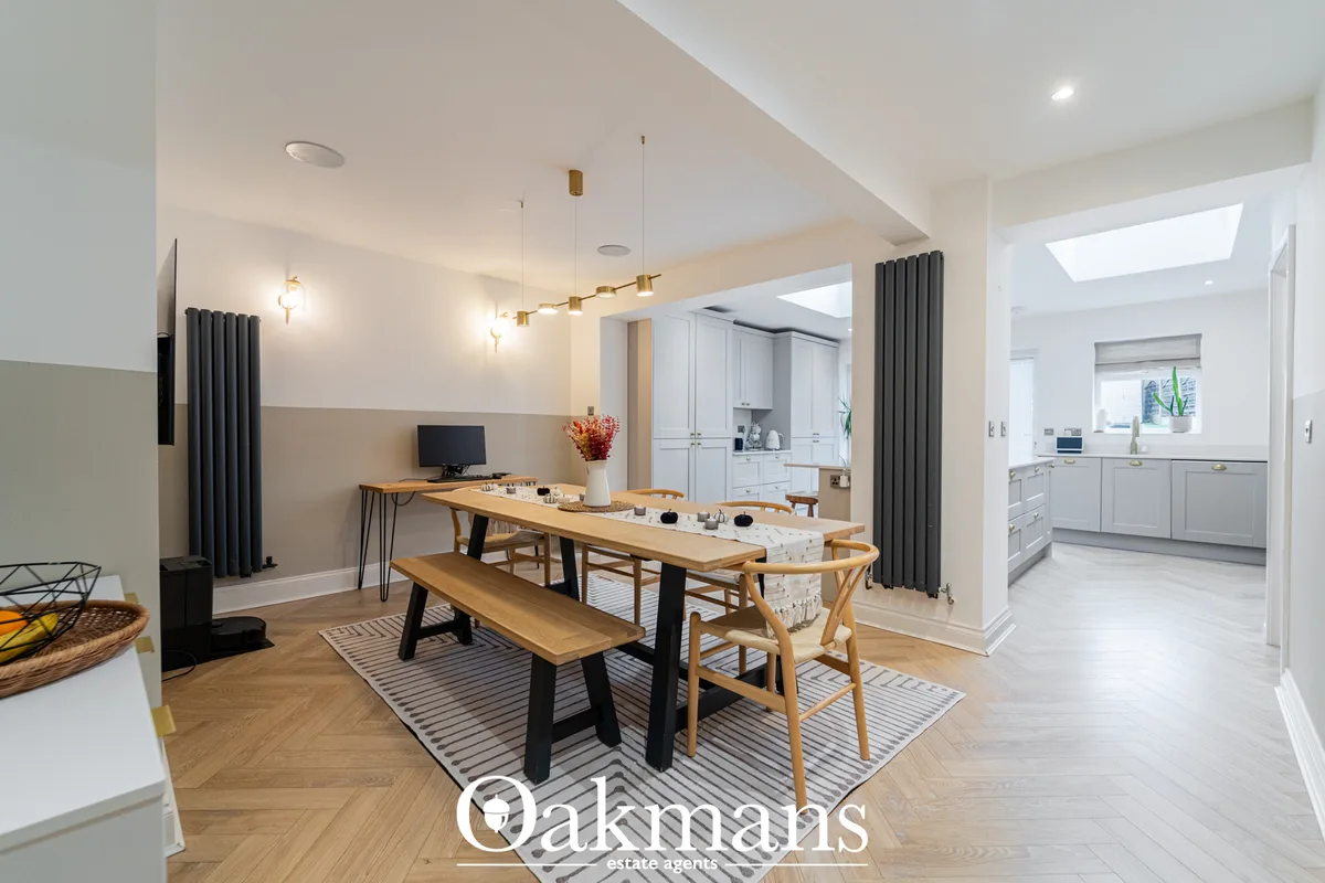 Open-plan dining area with wooden table, bench seating and chairs, leading to modern white kitchen with skylight