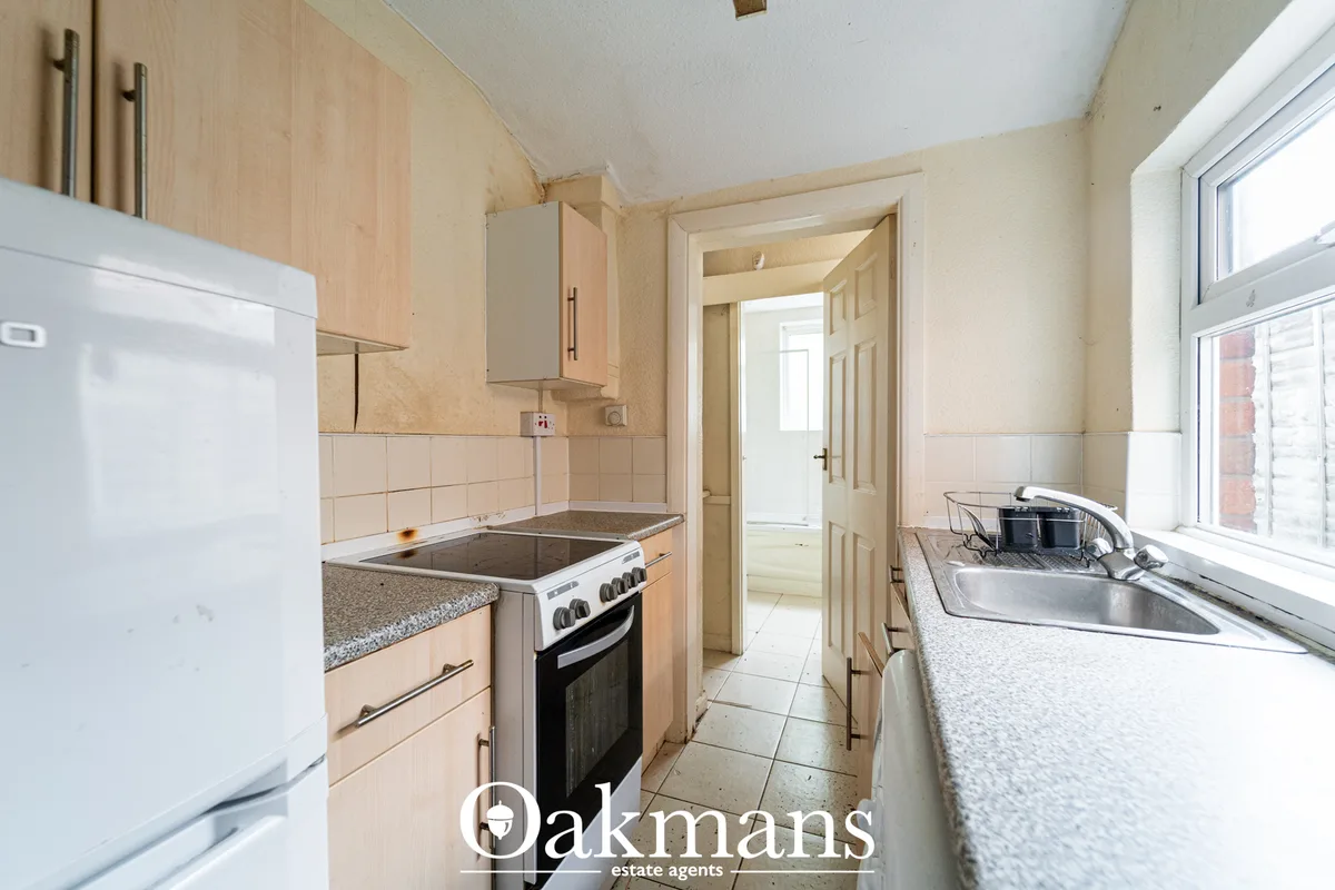 Galley kitchen with light wood units, granite worktops, electric oven, sink under window, cream tiles and white applianc