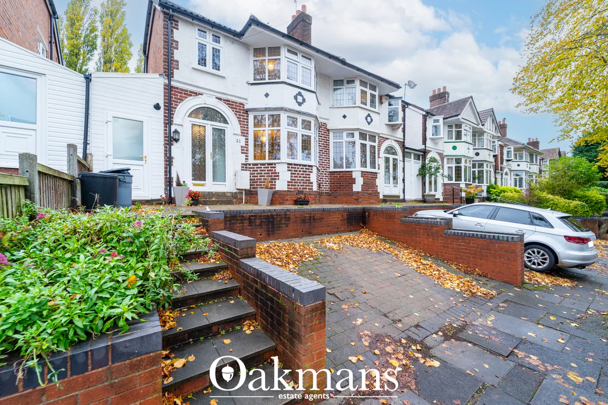 Red brick semi-detached house with white rendered gable, arched windows, front driveway and landscaped borders in autumn