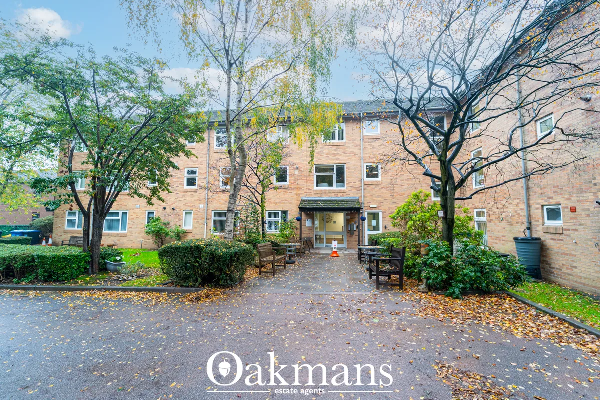 Brick residential building with central entrance, courtyard with autumn leaves, mature trees, benches and manicured hedg