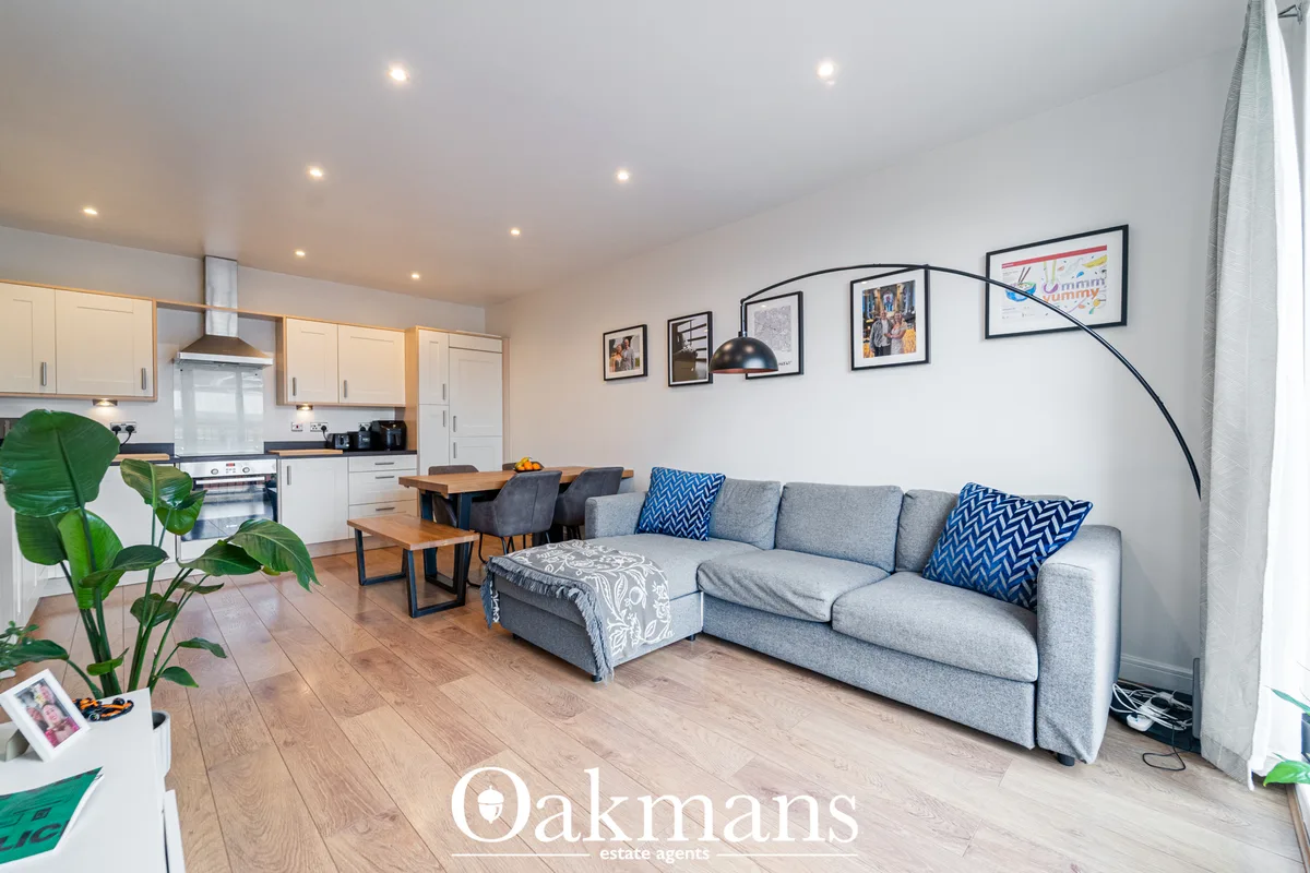 Open-plan living room with grey corner sofa, wooden flooring, framed wall art, arc floor lamp and access to fitted kitch